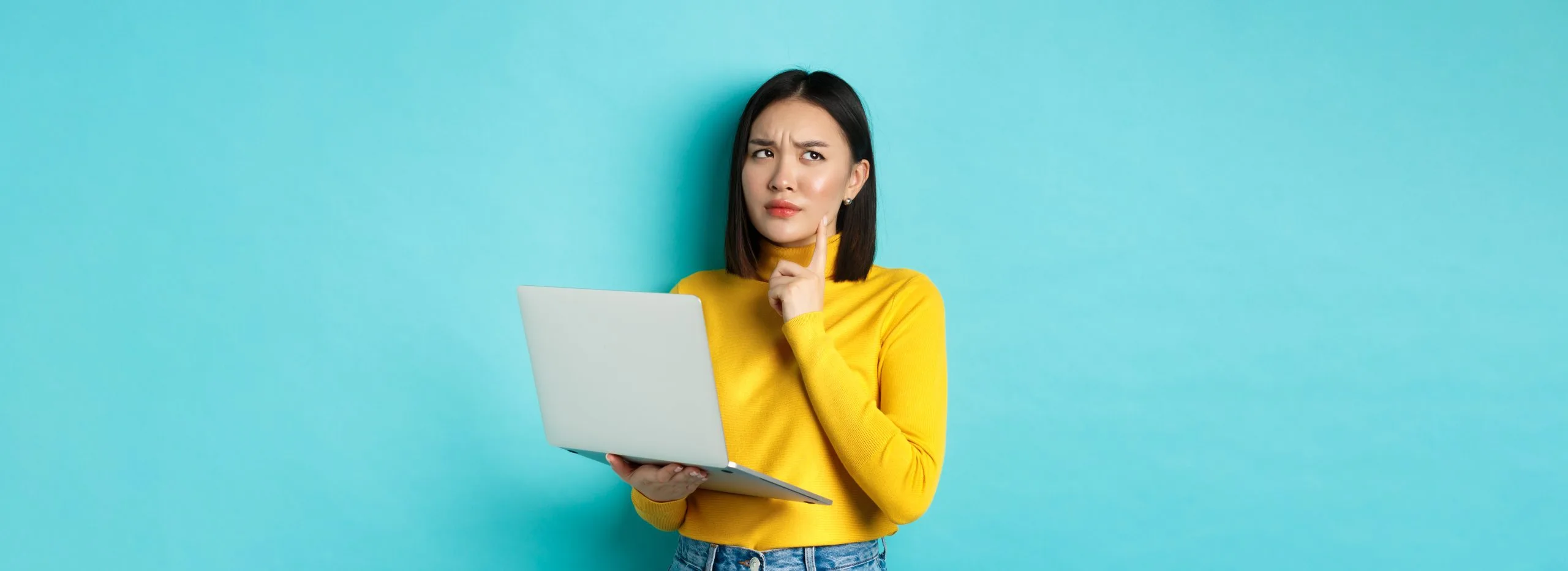 Pensive asian woman working on laptop, thinking and looking away, making decision, standing against blue background